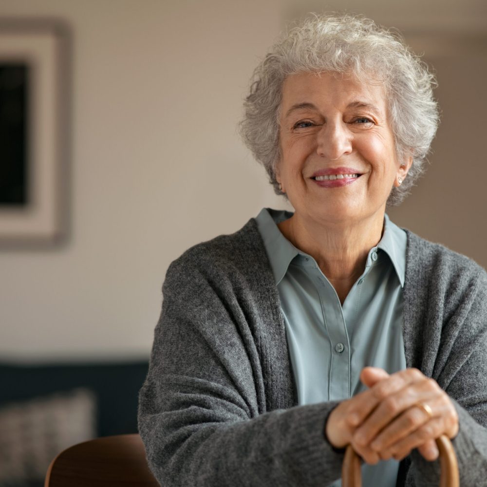 Portrait of smiling mature woman holding walking stick and sitting on chair at home. Portrait of happy senior woman under quarantine during covid-19 pandemic smiling while looking at camera. Old retired lady with grey and white hair stay at nursing home with copy space.